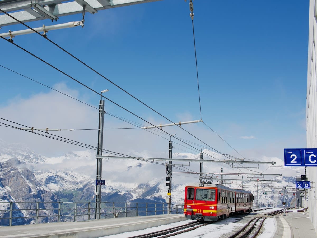Red train on snowy tracks with mountains under blue sky.